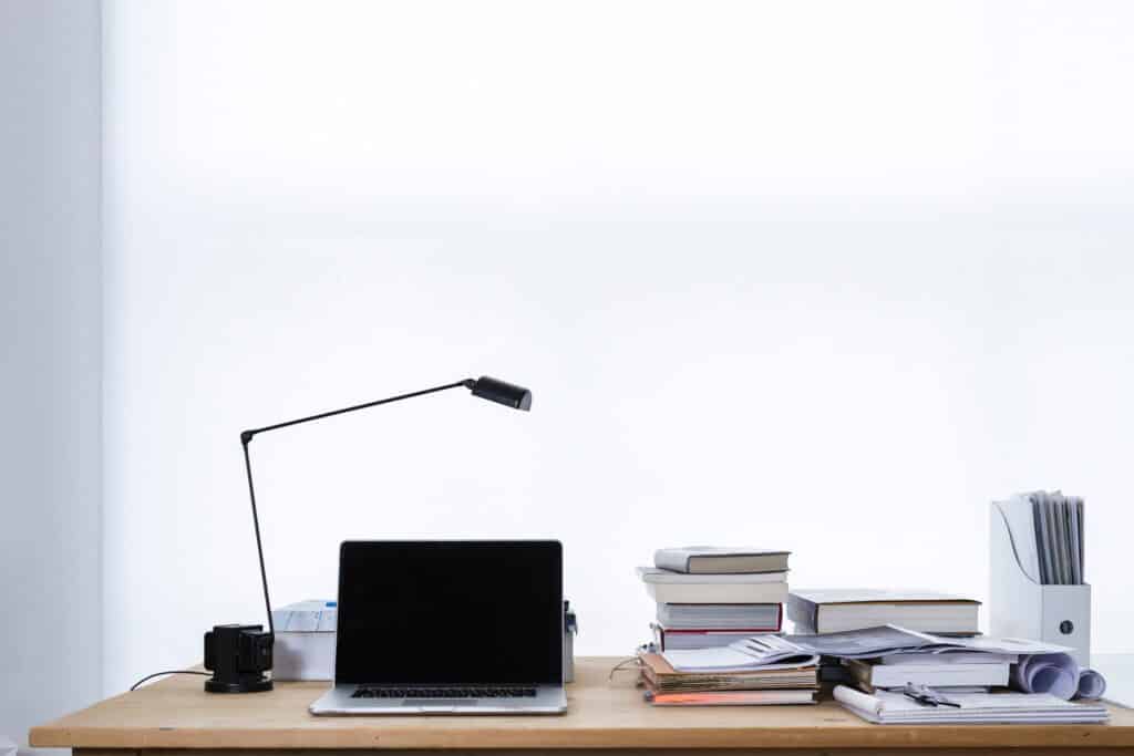 a wooden work desk with a laptop, a lamp shade, a file organiser, and stacks of books