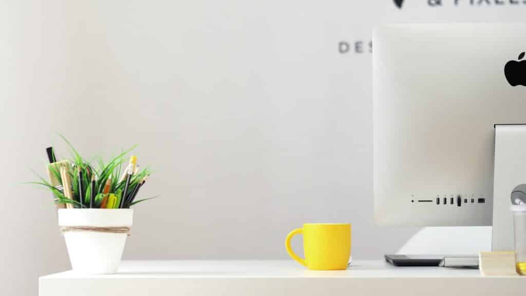 a white work desk showing the back of an apple dekstop, a yellow coffee mug an white flower vase