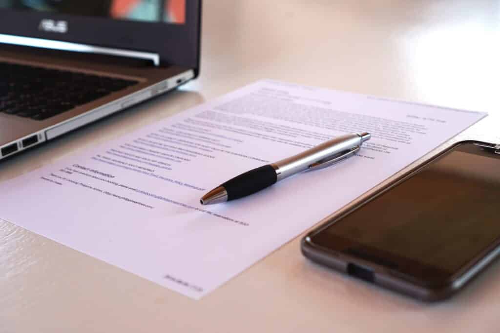 A printed contract placed on top of a table and between a laptop and a mobile phone.