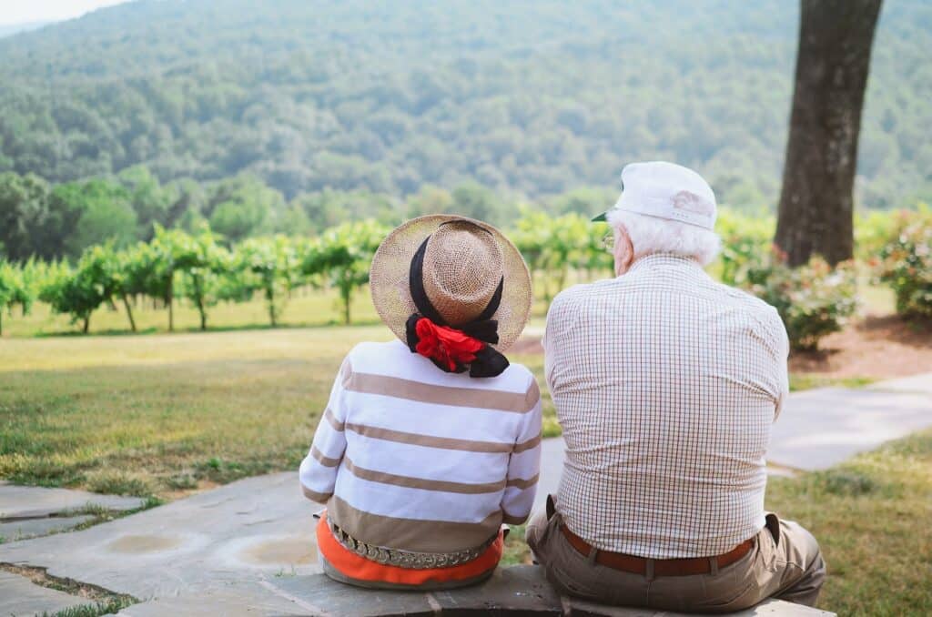 a retired couple sitting on a pathway at watching the trees and vineyard before them