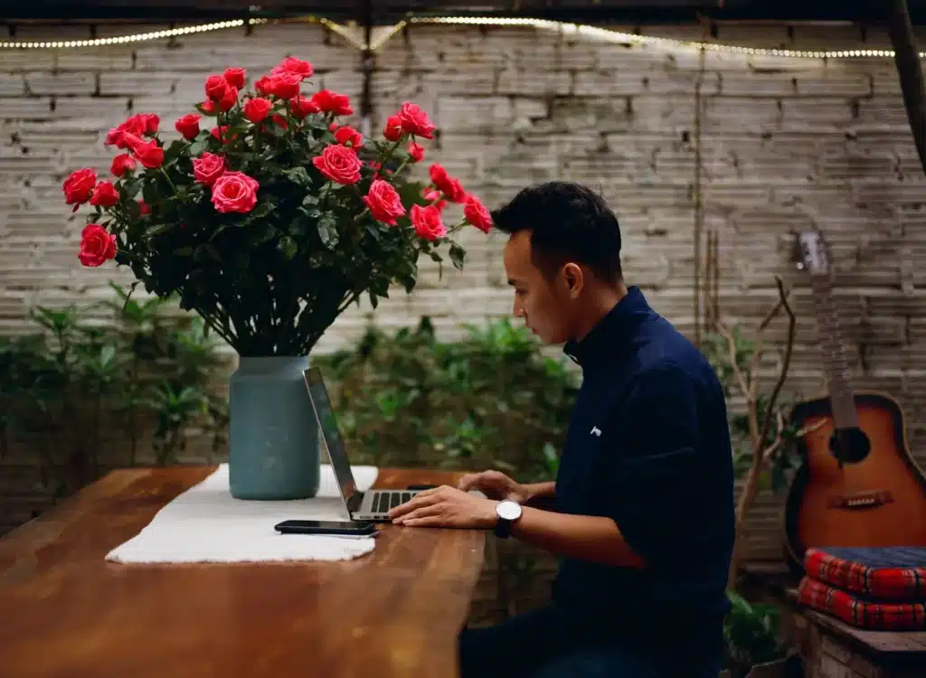 a young man in a restaurant, working on his laptop placed atop a mahogany table with a large vase filled with red roses
